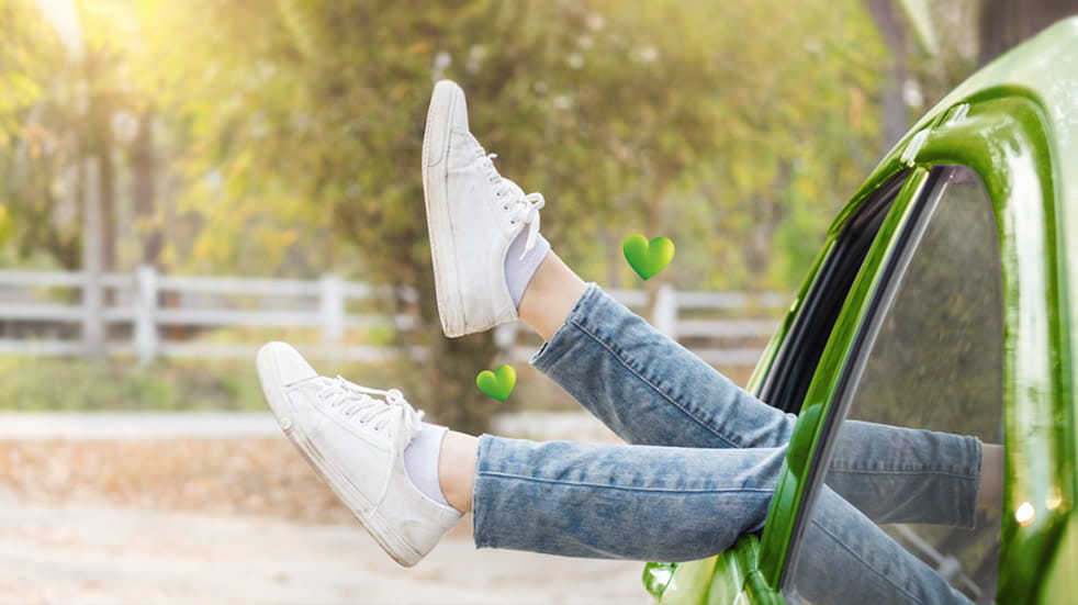 Green coloured car with window wound down, parked in outdoor space. Woman’s legs are sticking out of the window, she wears blue jeans and white trainers. Two green coloured hearts float around her ankles. 